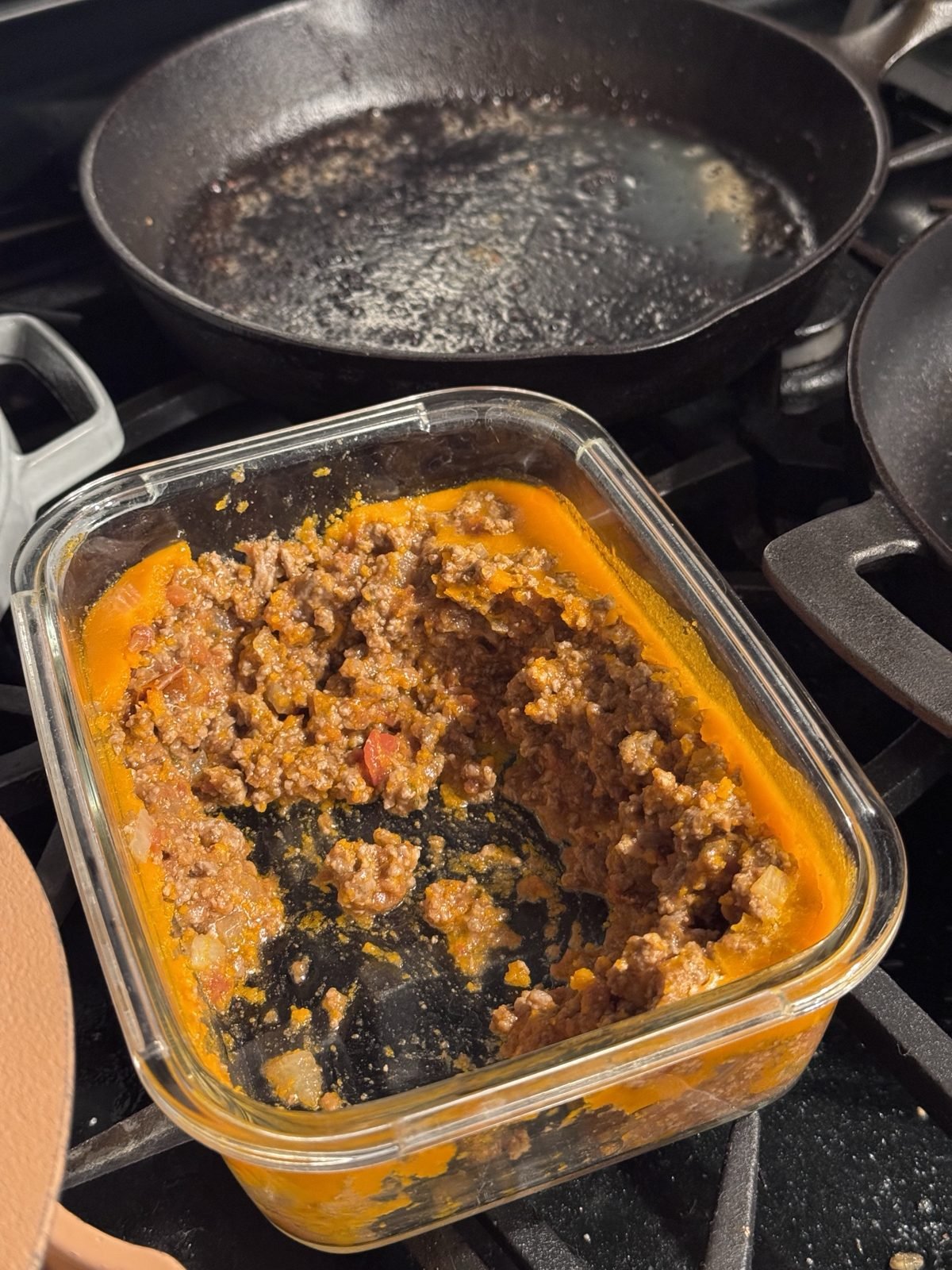 Glass container of ground beef in red sauce over baked potato, cast iron pan behind it
