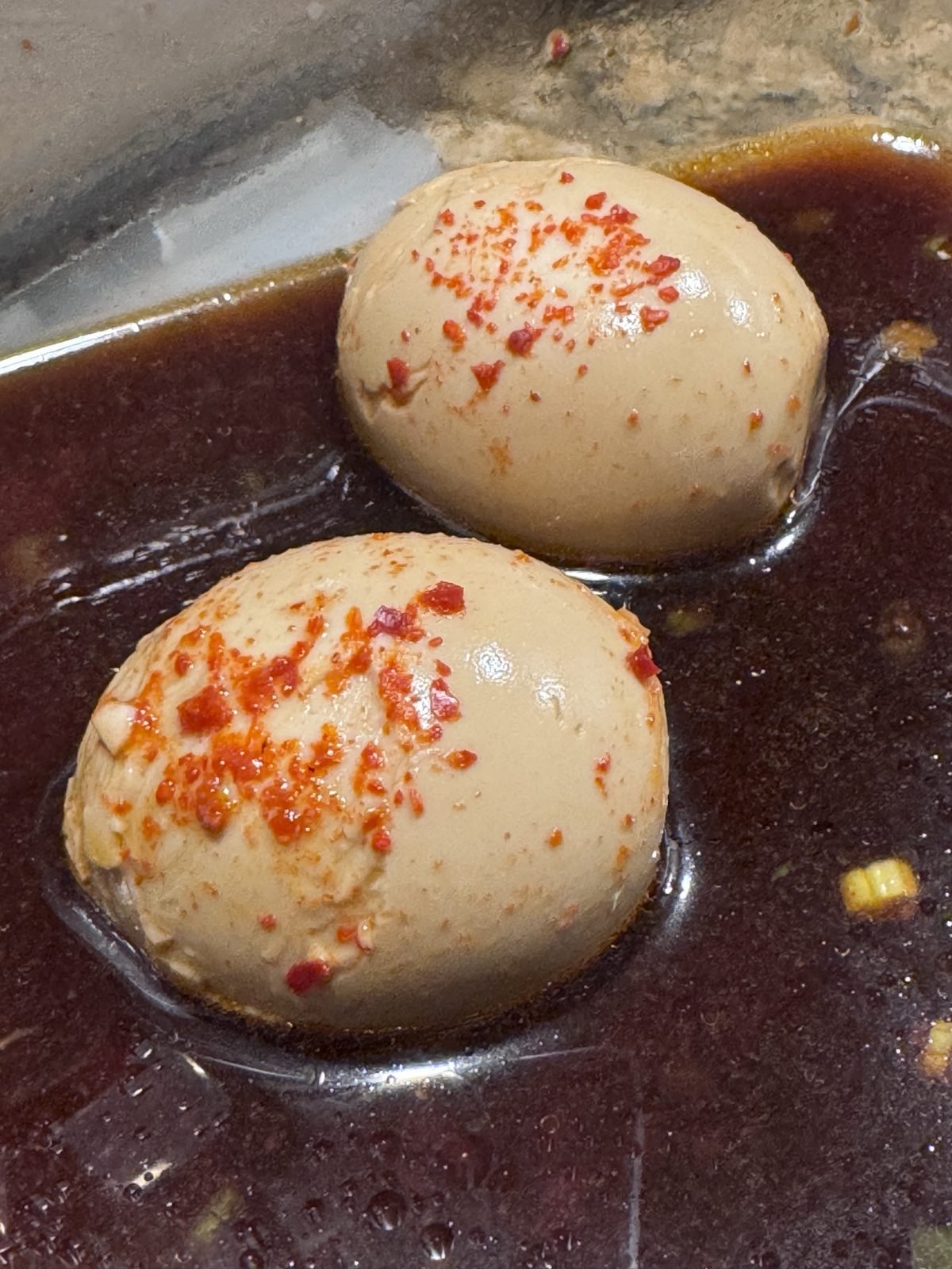 Two peeled mayak eggs soaking in dark soy marinade, topped with red chili flakes